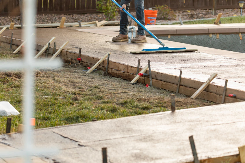 Worker smoothing freshly poured concrete on a backyard walkway using a large blue tool, with wooden framing visible.