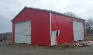 A large red metal building with two tall white garage doors and a single white entry door on a gravel lot.