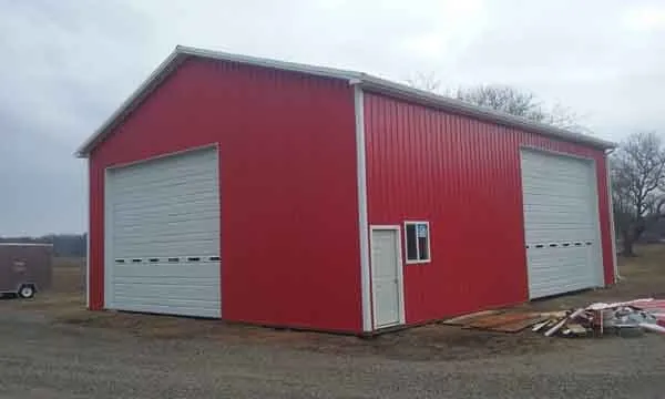 A large red metal building with two tall white garage doors and a single white entry door on a gravel lot.