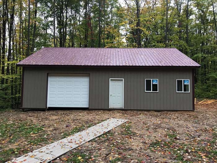 A brown metal-sided garage with a white door, garage door, and two windows, set against a wooded background.