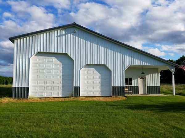 White metal barn with three large garage doors, a small entry door, and a covered porch, set on a grassy lawn.