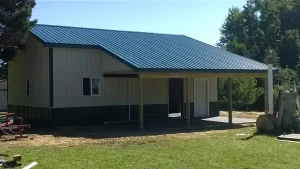 A small building with a blue metal roof and covered porch stands on a grassy lot with trees in the background.