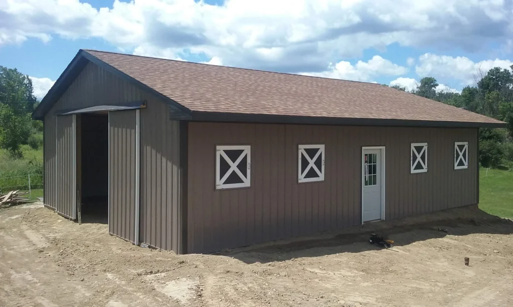 A brown metal barn with white-trimmed windows and a door sits on a dirt lot under a partly cloudy sky.
