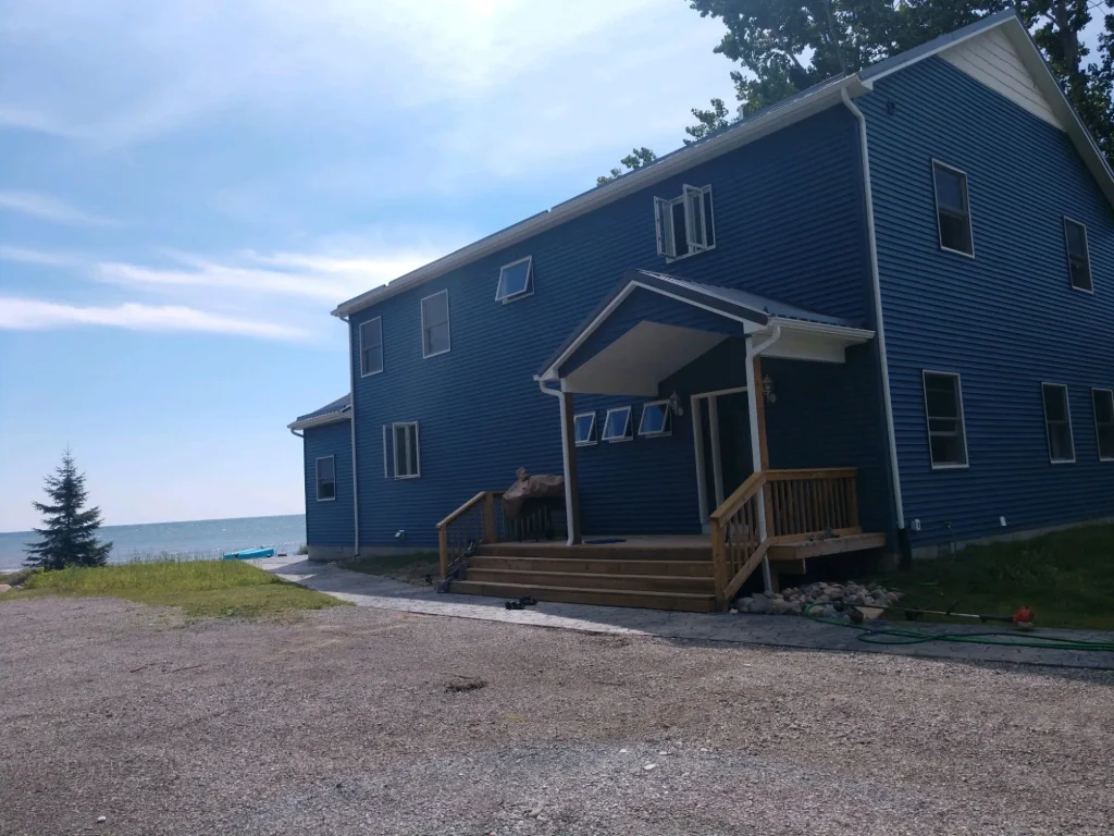 A blue two-story house with a front porch sits near a gravel driveway and overlooks a body of water.