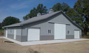 A large gray metal building with three white garage doors and a concrete driveway, surrounded by trees.
