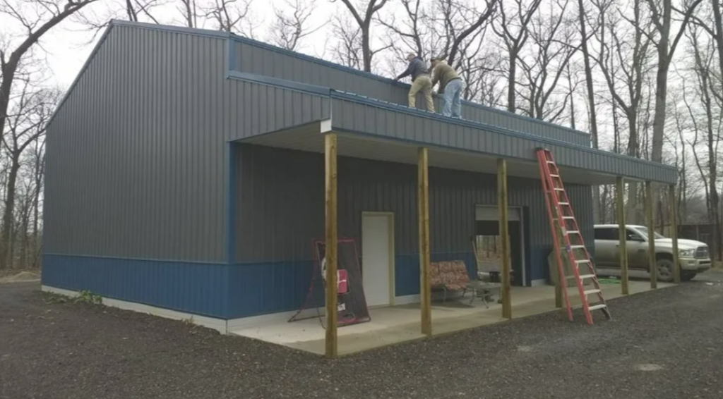 Two people work on the roof of a blue metal building with a porch; a red ladder leans against the front.